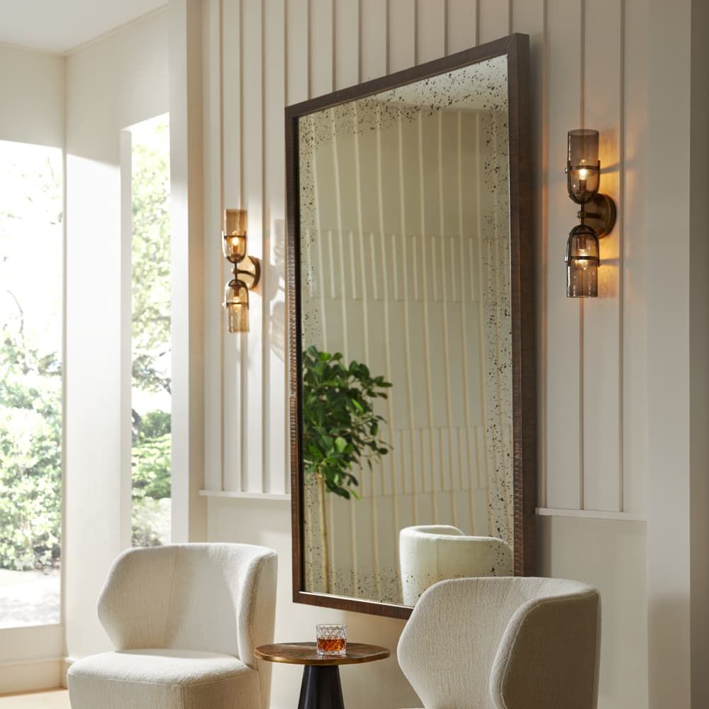 Large mirror on a wall with two chairs and a small table in a room with large windows.