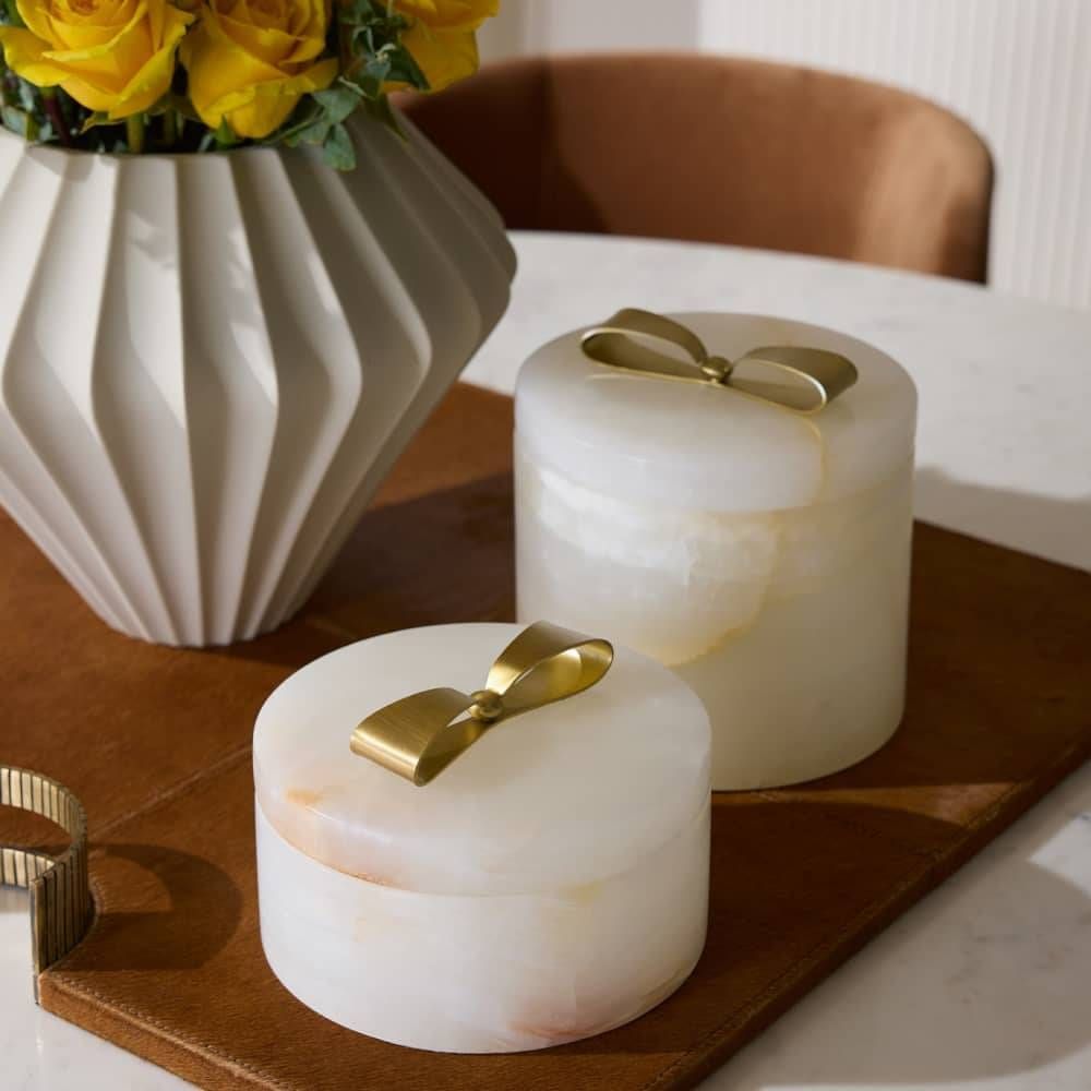 Two marble jars with gold lids on a wooden tray next to a vase with yellow flowers.