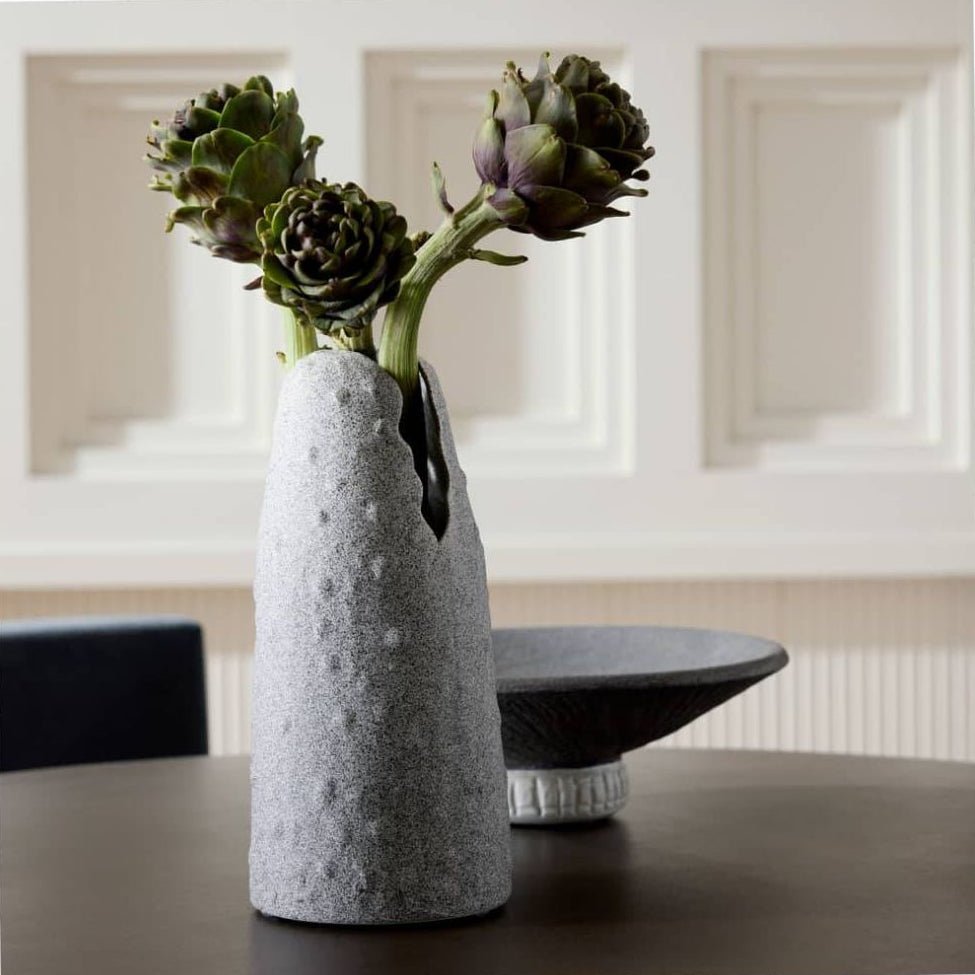 Gray textured vase with artichokes on a table in a room with white walls.