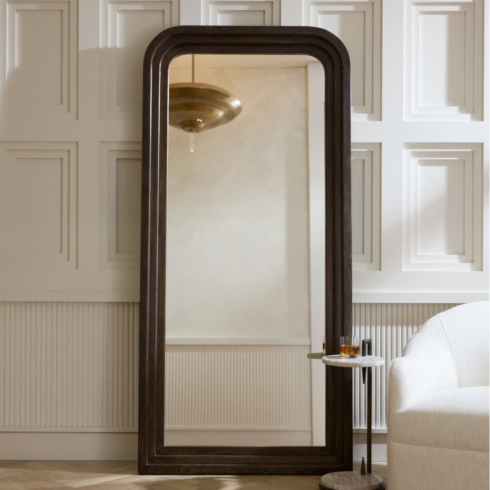 Large floor mirror with dark wood frame in a room with white paneled walls and a white sofa.