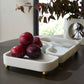 White decorative tray with plums on a dark surface, blurred plant in the background