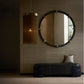 A modern interior scene featuring a large round wall mirror with a dark frame and brass accents, a cylindrical pendant light with hanging metallic strands, and a dark upholstered bench against a textured beige wall.