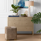 Beige dresser with decorative items and a lamp, featuring a plant and abstract art on a light-colored wall.