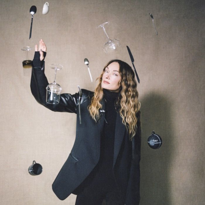 Person in a black outfit with hanging glasses, spoons, and other objects against a beige wall.