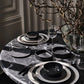 Dining table setting with black and white ceramic plates, glasses, and cutlery on a marble surface.