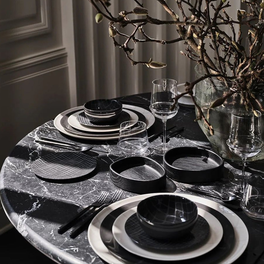 Dining table setting with black and white ceramic plates, glasses, and cutlery on a marble surface.
