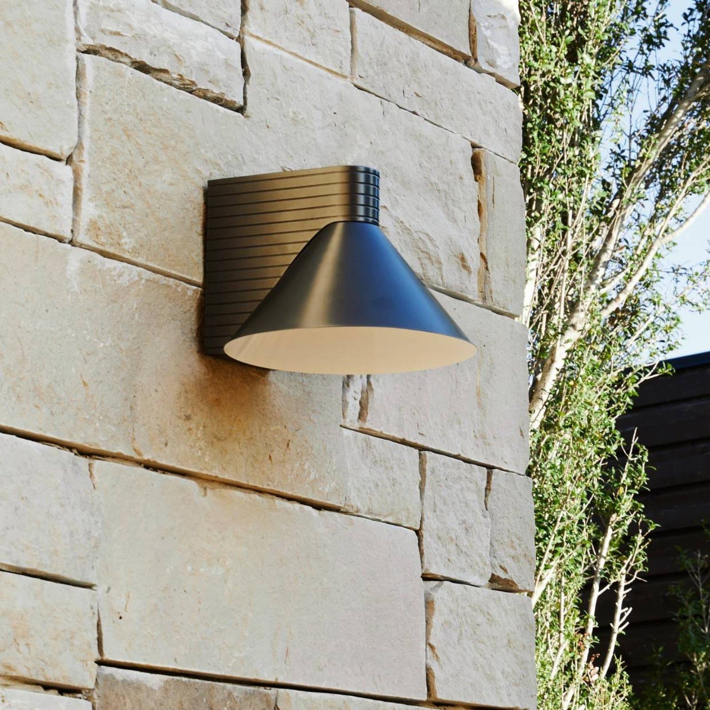 Modern outdoor wall light fixture on a stone wall with greenery in the background