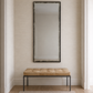 A tall rectangular wall mirror with a dark distressed frame hangs above a tufted bench with a tan cushion and black metal legs, set against a textured beige wall in a bright entryway.