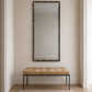 A tall rectangular wall mirror with a dark distressed frame hangs above a tufted bench with a tan cushion and black metal legs, set against a textured beige wall in a bright entryway.