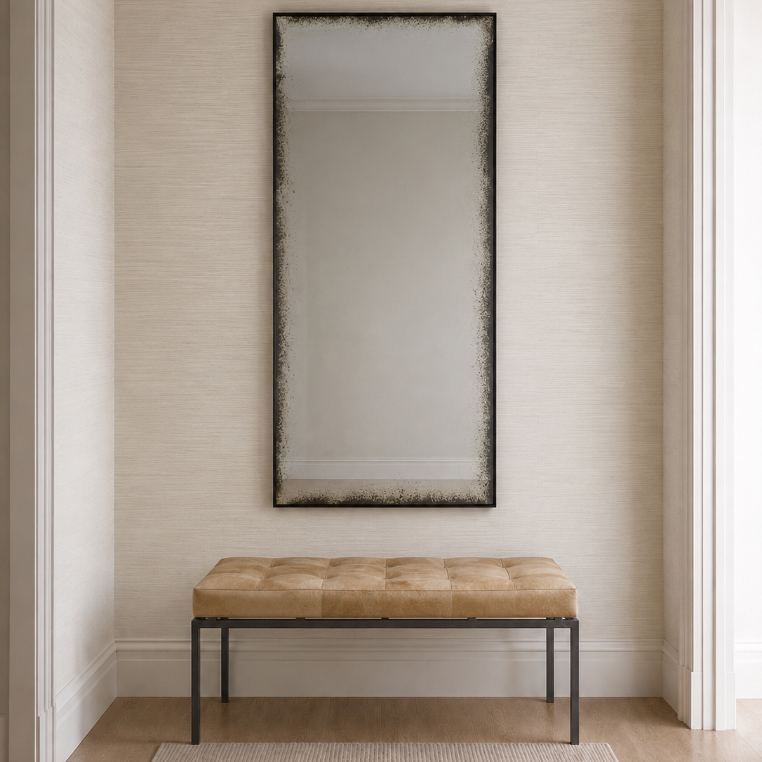 A tall rectangular wall mirror with a dark distressed frame hangs above a tufted bench with a tan cushion and black metal legs, set against a textured beige wall in a bright entryway.