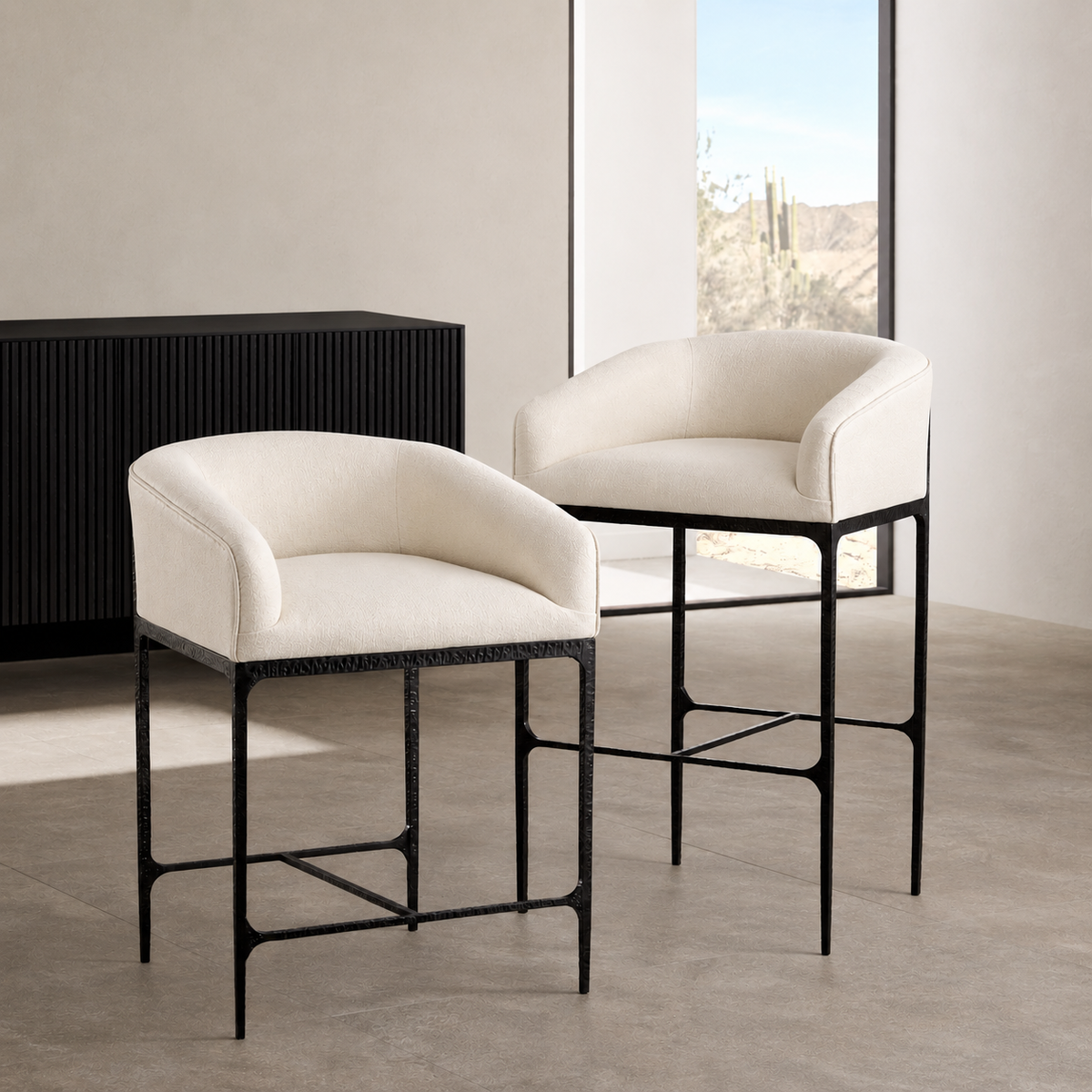 Two upholstered bar stools with cream fabric seats and backs and black metal frames, shown in a modern interior setting with a window and concrete floor.