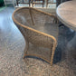 A woven rattan accent chair with a curved back and seat, positioned on a speckled terrazzo floor next to a round table.