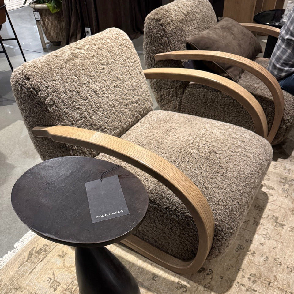 Gray upholstered chair with wooden armrests and a small round dark table on a patterned rug.
