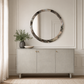 A round wall mirror with a textured, marbled gray and brown frame hangs above a light gray sideboard in a styled room setting with a plant and bowl.