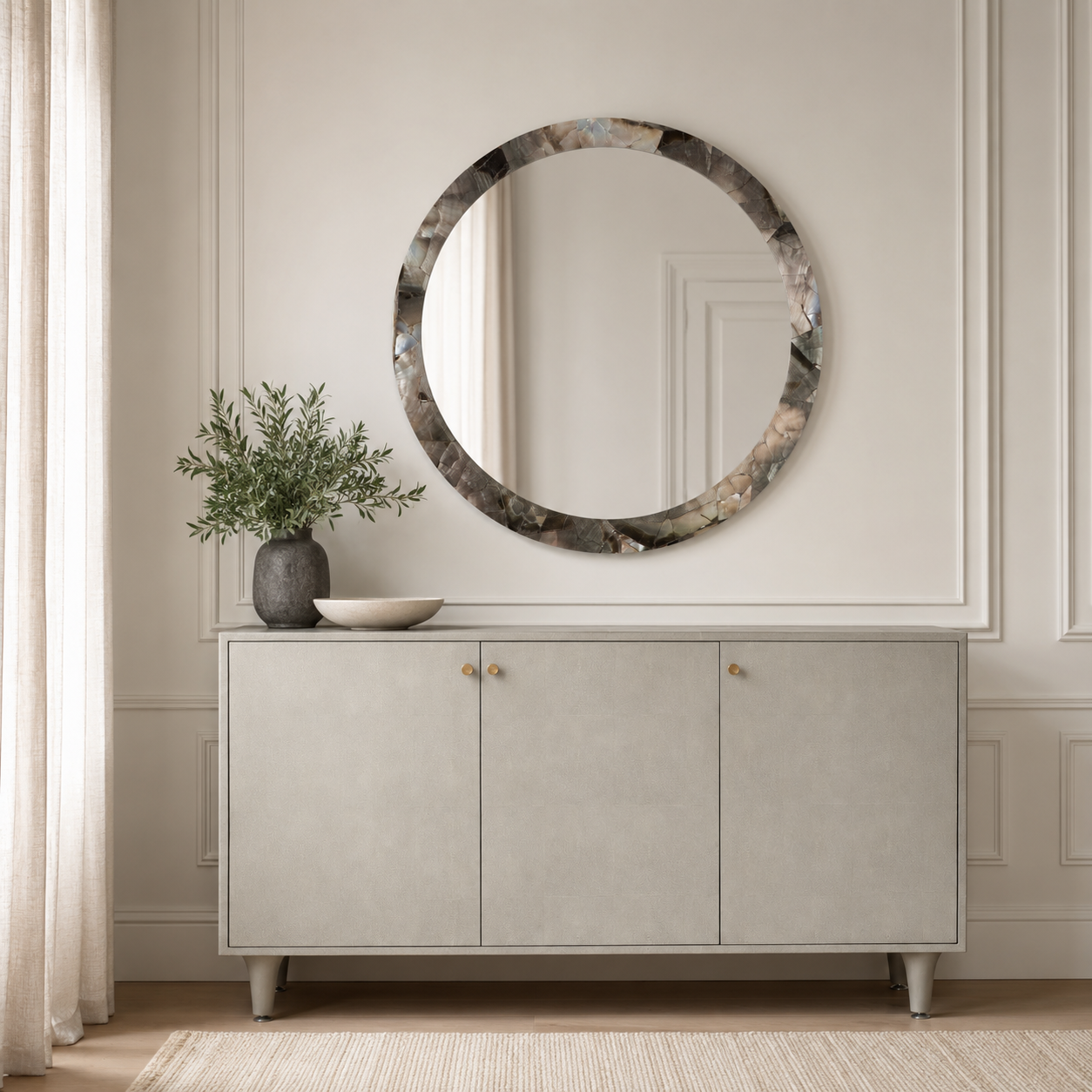 A round wall mirror with a textured, marbled gray and brown frame hangs above a light gray sideboard in a styled room setting with a plant and bowl.