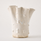 An organic-shaped ceramic vase with a flared, wavy rim and a glossy, textured surface featuring bubble-like patterns, in a warm cream or off-white color, set against a plain light background.