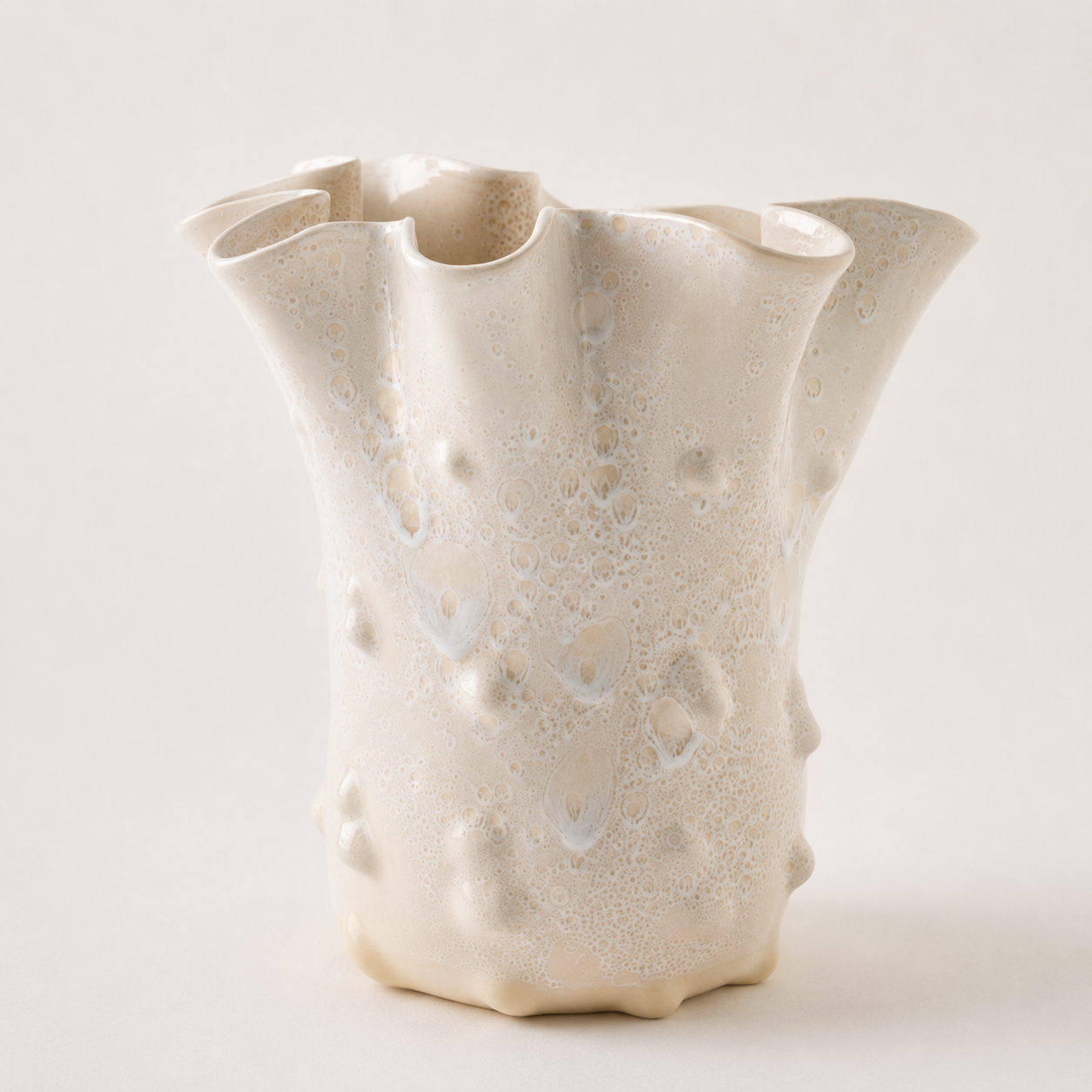 An organic-shaped ceramic vase with a textured, bubbly surface and flared, wavy rim, in a warm cream or off-white glaze against a light neutral background.