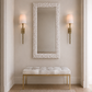 A tall rectangular wall mirror with a textured white frame hangs above a tufted bench with a white cushion and gold legs in a neutral entryway. Two gold sconces with white shades flank the mirror on beige walls.