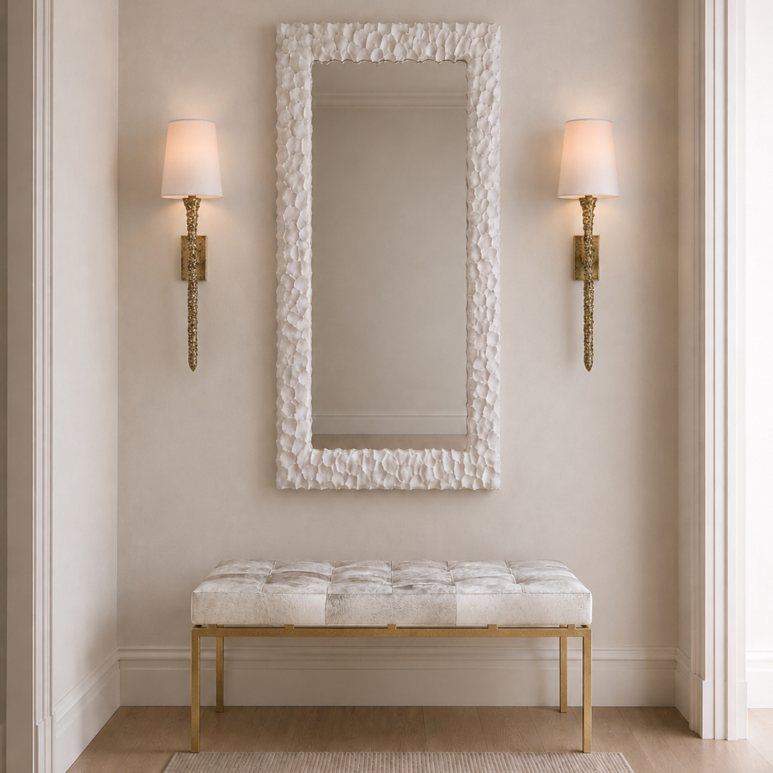 A tall rectangular wall mirror with a textured white frame hangs above a tufted bench with a white cushion and gold legs in a neutral entryway. Two gold sconces with white shades flank the mirror on beige walls.