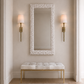A tall rectangular wall mirror with a textured white frame hangs above a tufted bench with a white cushion and gold legs in a neutral entryway. Two gold sconces with white shades flank the mirror on beige walls.