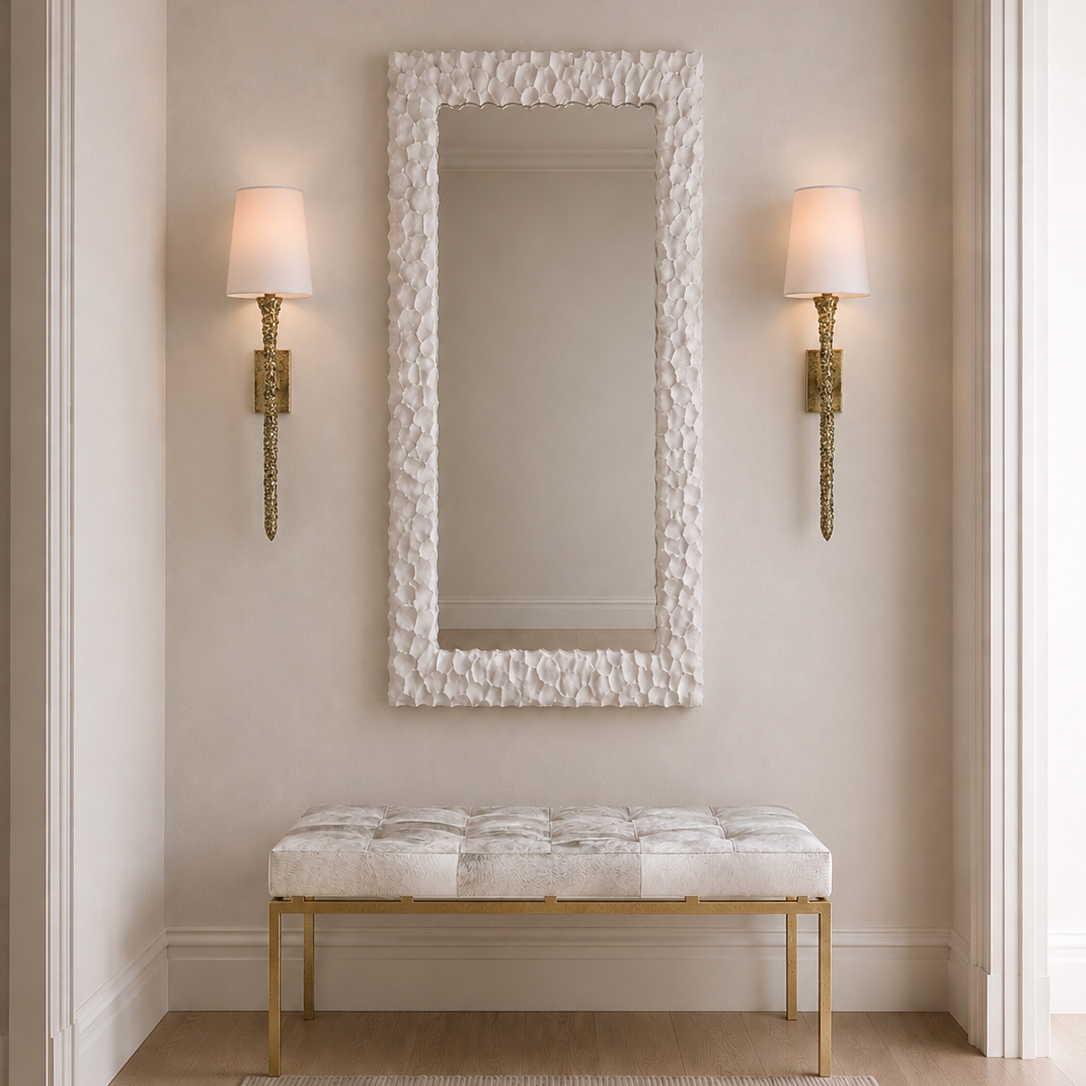 A tall rectangular wall mirror with a textured white frame hangs above a tufted bench with a white cushion and gold legs in a neutral entryway. Two gold sconces with white shades flank the mirror on beige walls.