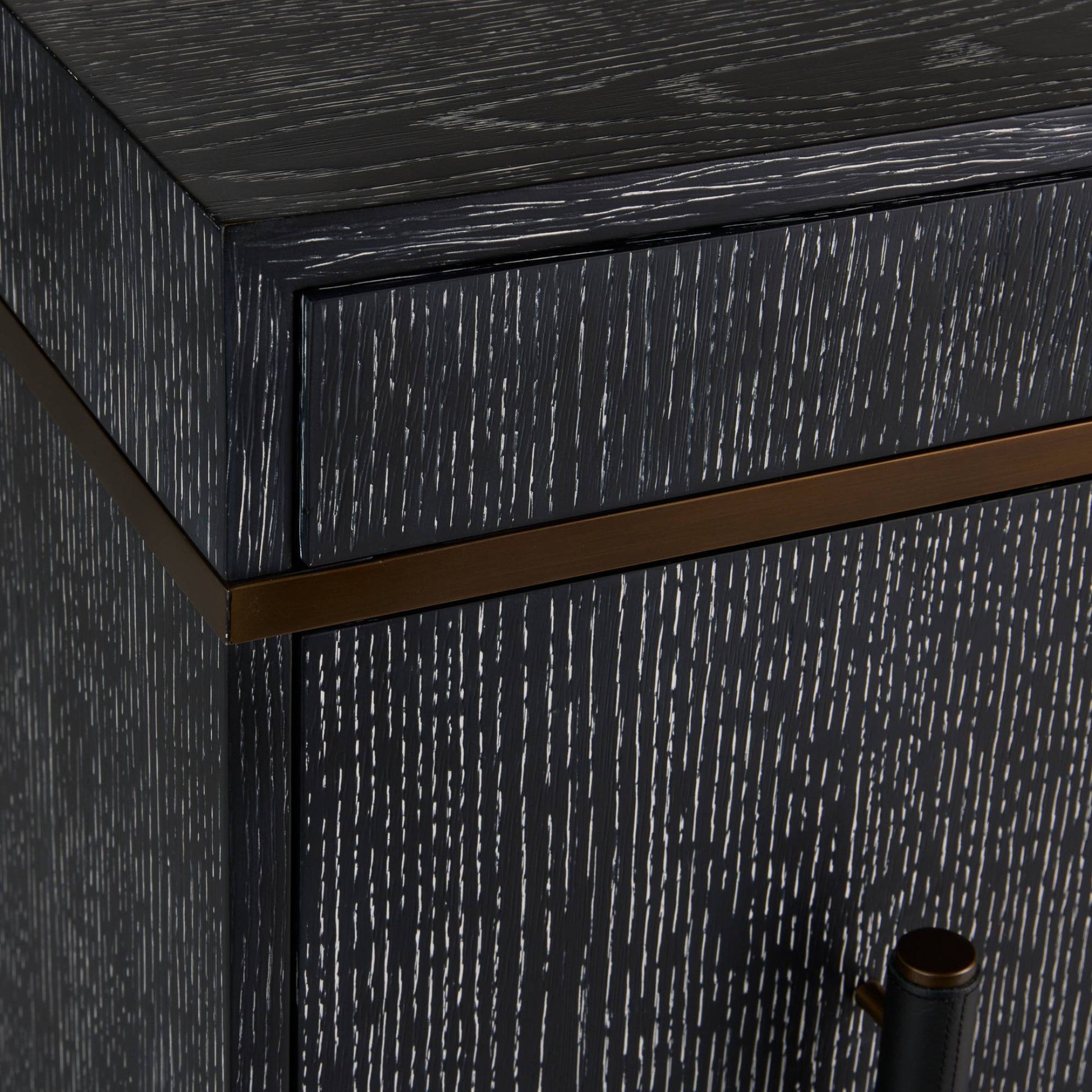 Close-up of a dark wood cabinet corner with a distressed black finish, a horizontal bronze metal accent strip, and part of a dark metal handle visible.