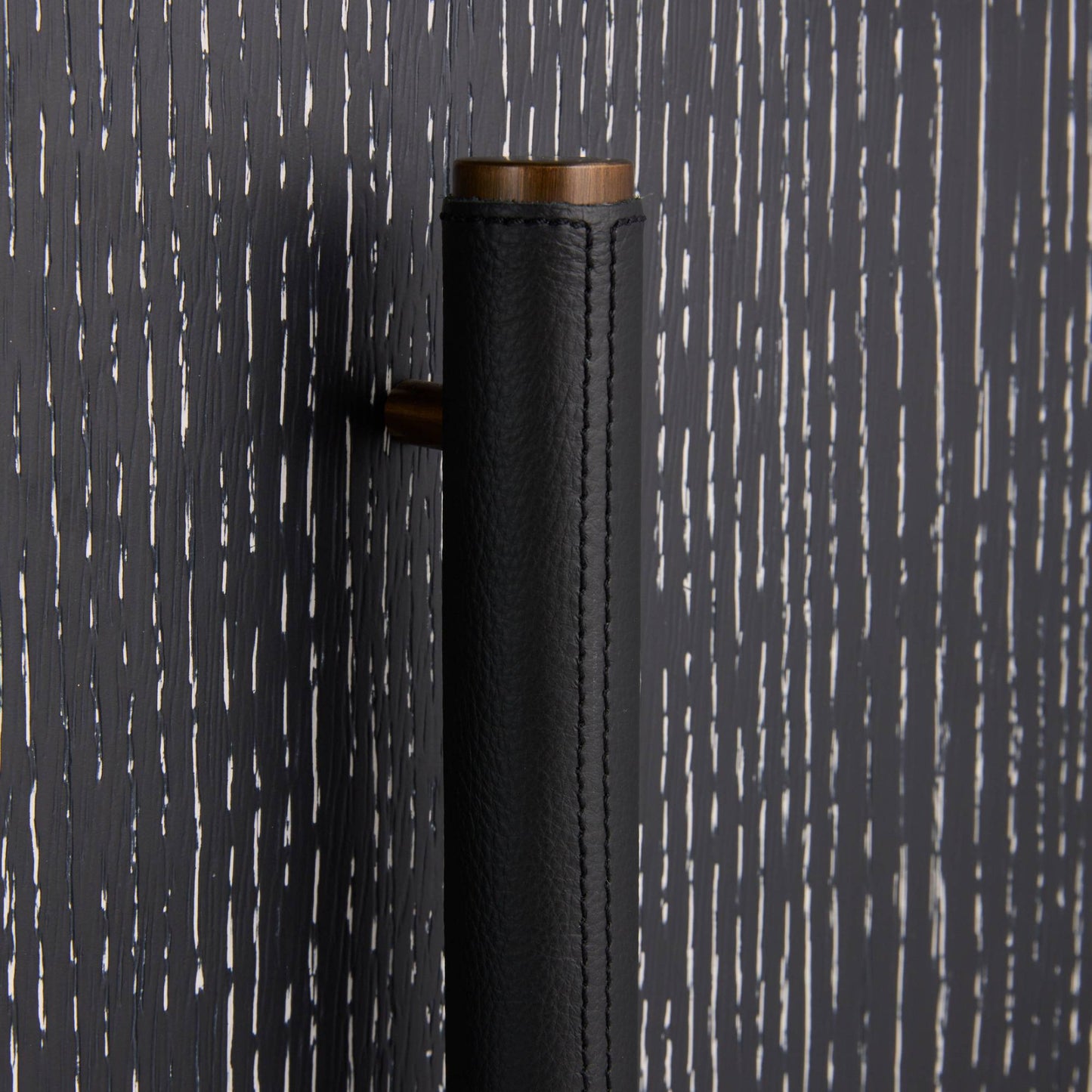 A close-up of a modern cylindrical door handle featuring a textured black leather wrap and a warm bronze metal top and side hardware, set against a dark charcoal wood-grain background.