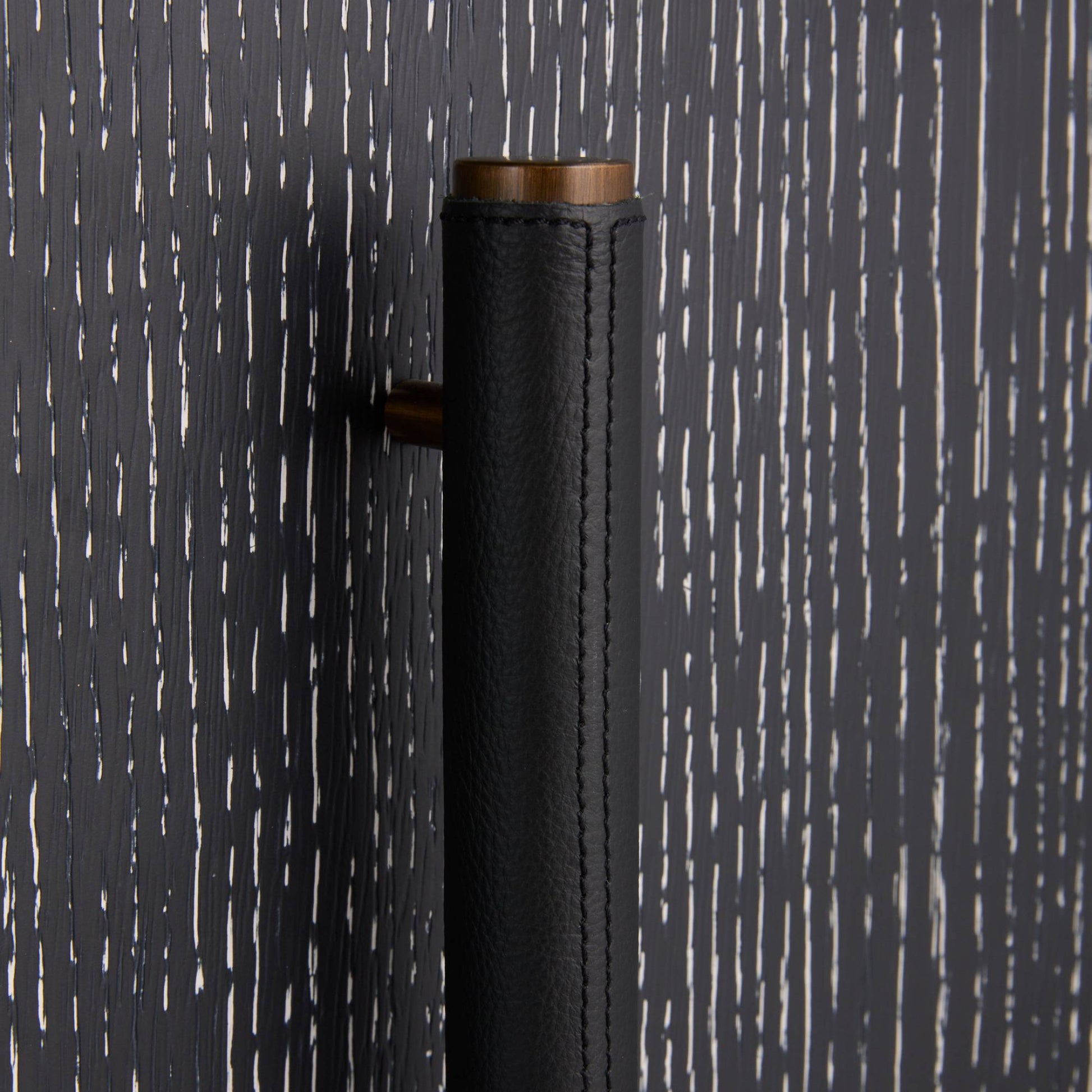A close-up of a modern cylindrical door handle featuring a textured black leather wrap and a warm bronze metal top and side hardware, set against a dark charcoal wood-grain background.