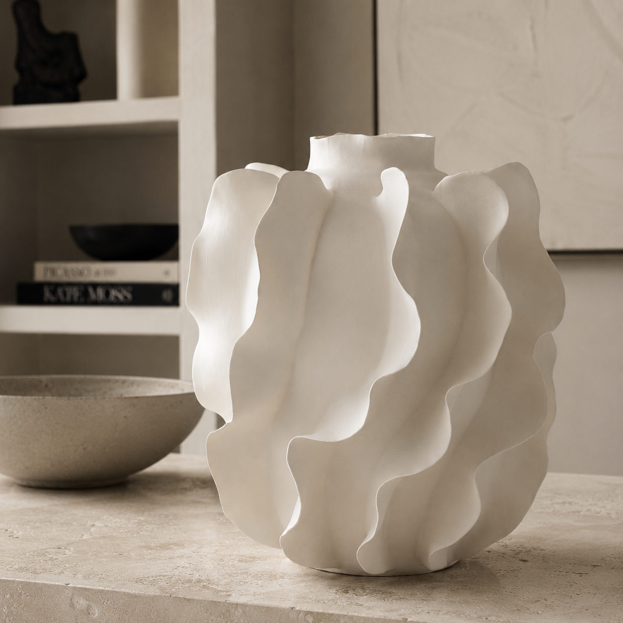 A white ceramic vase with a wavy, organic ribbed texture, displayed on a stone surface next to a textured bowl with a blurred shelving unit in the background.