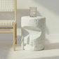 A white ceramic side table with a textured, speckled finish and an abstract sculptural design, styled in a bright room next to a wooden chair with a woven seat