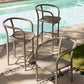 Three outdoor bar stools with curved taupe metal frames and woven seats, arranged on a patio beside a swimming pool.