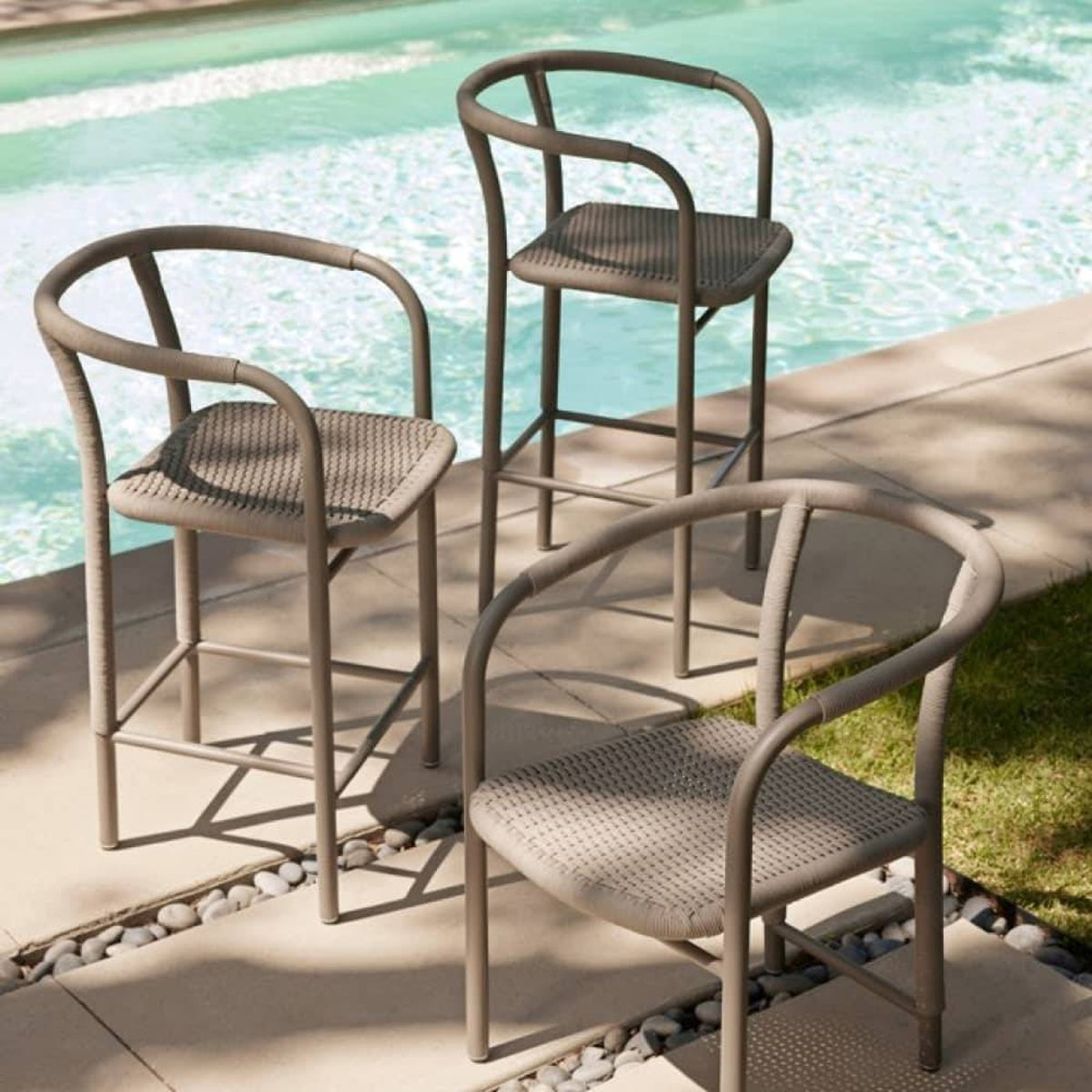 Three outdoor bar stools with curved taupe metal frames and woven seats, arranged on a patio beside a swimming pool.