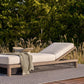 Outdoor lounge chair with white cushions and a green blanket on a patio with grass and trees in the background.