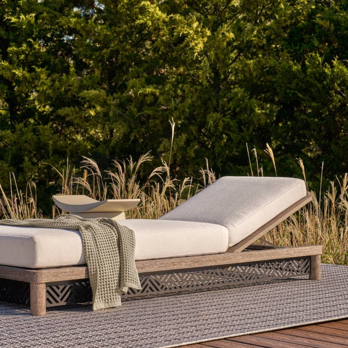 Outdoor lounge chair with white cushions and a green blanket on a patio with grass and trees in the background.
