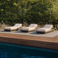 Three outdoor chaise lounge chairs with cream cushions and light wood frames arranged on a slatted deck beside a swimming pool, with tall grass and dense green trees in the background.