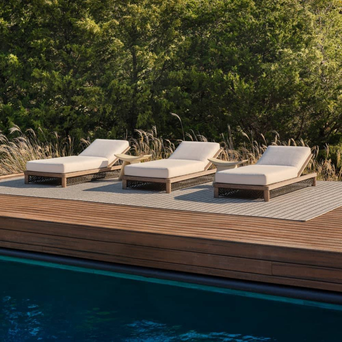 Three outdoor chaise lounge chairs with cream cushions and light wood frames arranged on a slatted deck beside a swimming pool, with tall grass and dense green trees in the background.