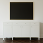 An off-white wooden sideboard or credenza with three doors and drawers, styled in a warm interior setting with two matching table lamps and a framed landscape painting on a beige wall.