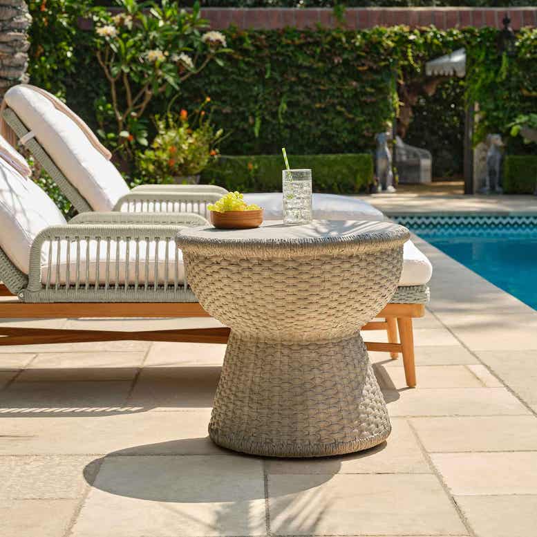 Wicker table with a drink and fruit bowl next to lounge chairs by a pool