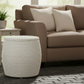 A round, ribbed white woven ottoman stool placed in front of a beige sofa with two cream pillows on a dark brick floor.