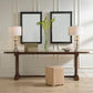 Console table with two lamps, a vase with greenery, and a small wooden box against a wall with framed mirrors.
