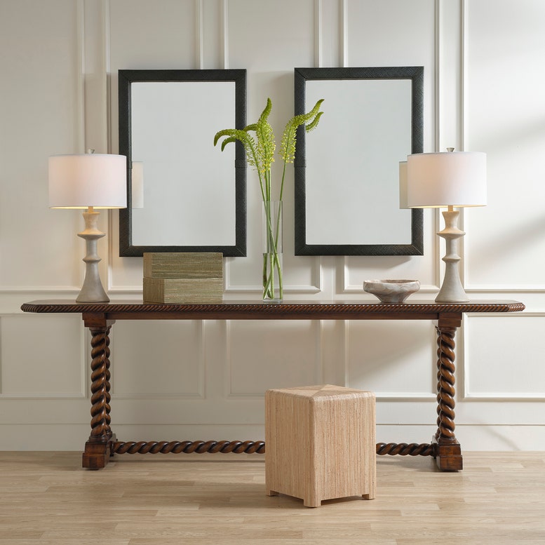 Console table with two lamps, a vase with greenery, and a small wooden box against a wall with framed mirrors.