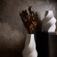 White vase with dried plants and a white abstract sculpture against a textured wall.