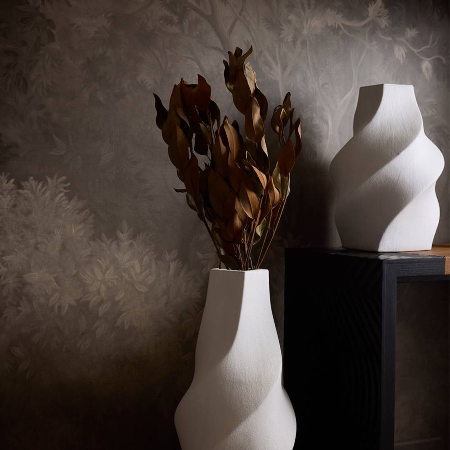 White vase with dried plants and a white abstract sculpture against a textured wall.