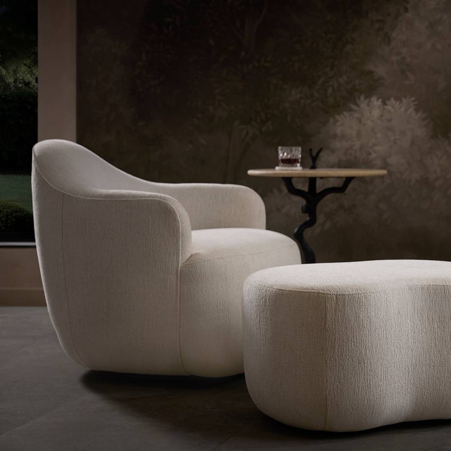 Modern beige armchair and ottoman with a small round table in a dimly lit room.