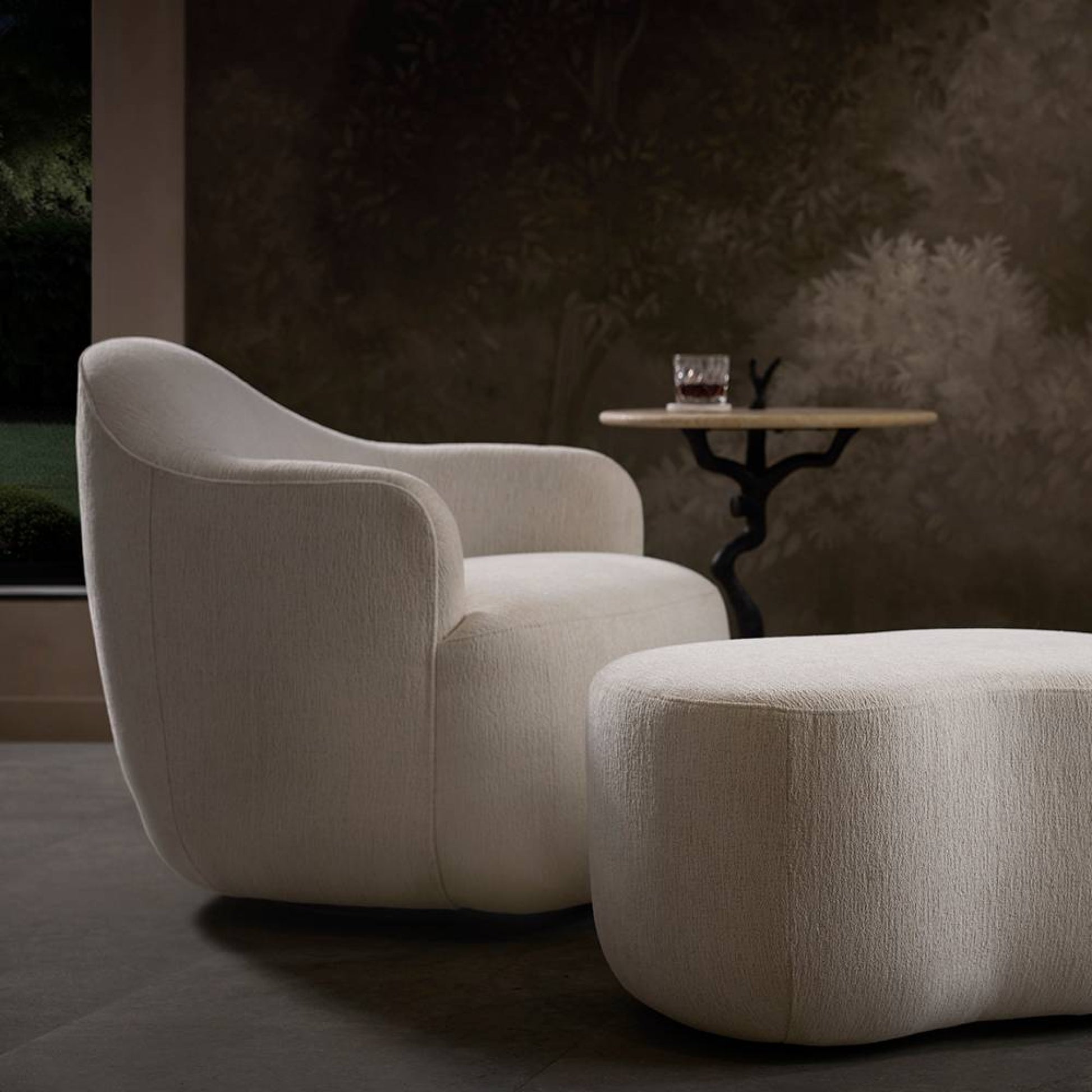 Modern beige armchair and ottoman with a small round table in a dimly lit room.