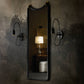 A tall rectangular wall mirror with a black frame hangs on a dark patterned wallpapered wall, flanked by two black wall sconces. The mirror reflects a softly lit interior scene with a table lamp and a white stool.