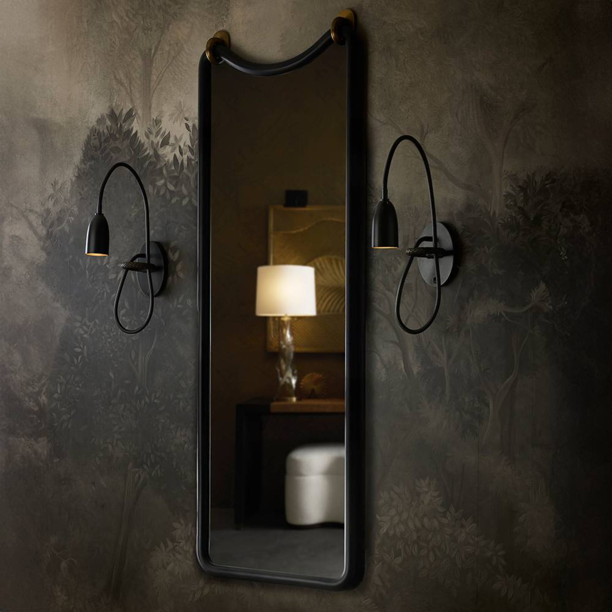 A tall rectangular wall mirror with a black frame hangs on a dark patterned wallpapered wall, flanked by two black wall sconces. The mirror reflects a softly lit interior scene with a table lamp and a white stool.