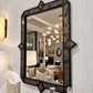 A large rectangular wall mirror with an ornate black frame featuring geometric corner details and a marbled dark pattern, mounted on a white textured wall. The mirror reflects a modern living room interior with a chandelier and sofa.