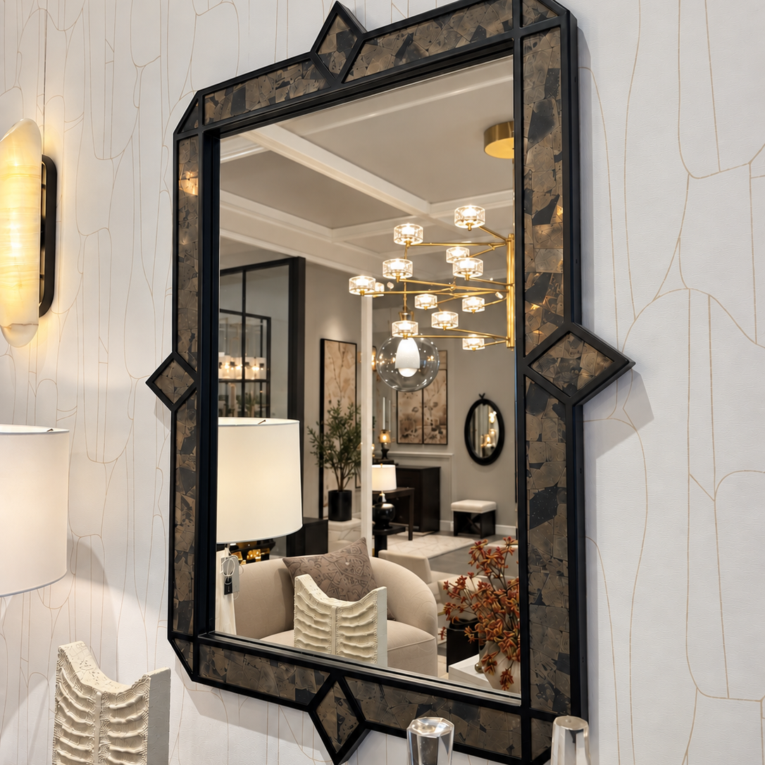 A large rectangular wall mirror with an ornate black frame featuring geometric corner details and a marbled dark pattern, mounted on a white textured wall. The mirror reflects a modern living room interior with a chandelier and sofa.