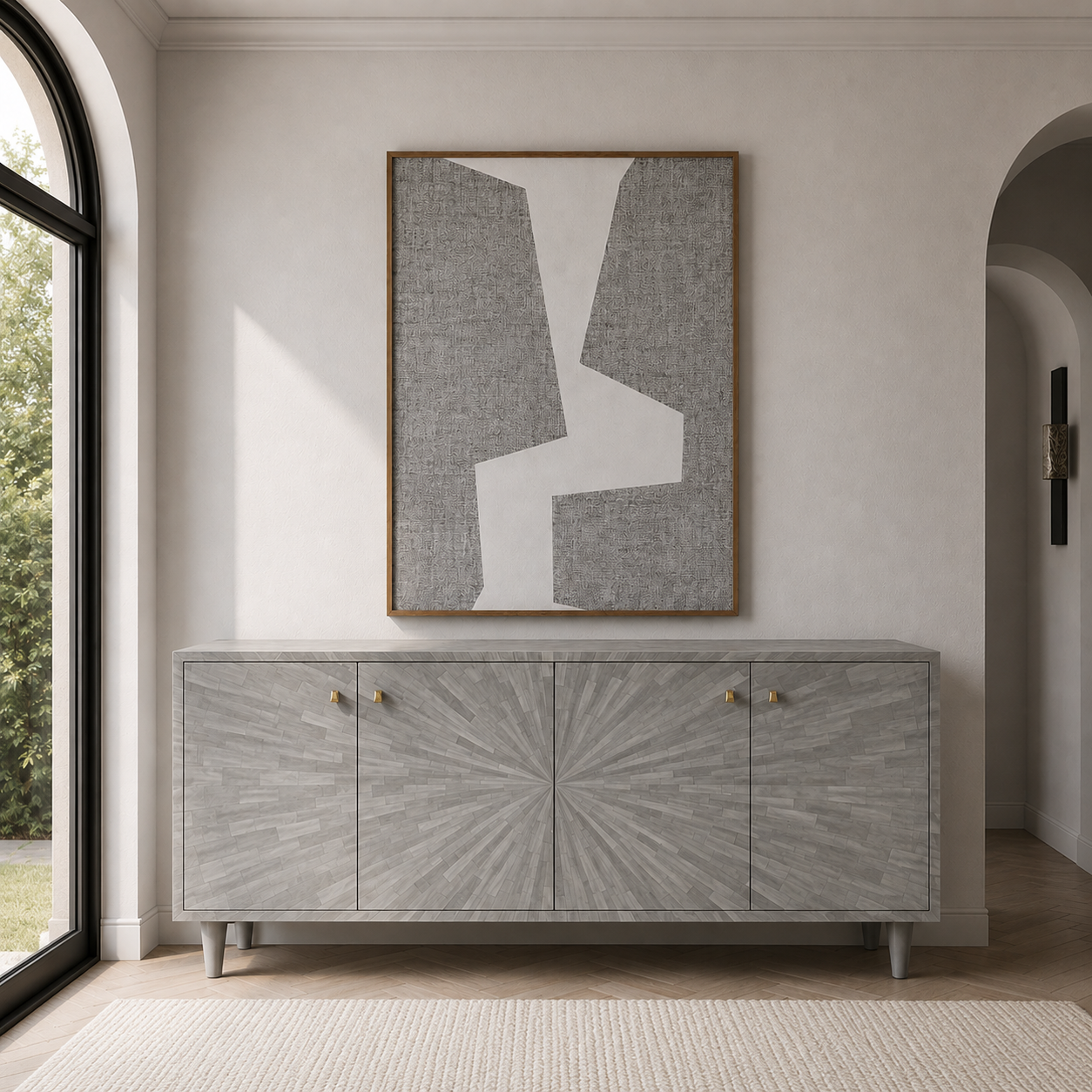 A modern gray wood-look sideboard cabinet with a sunburst center panel and brass knobs, styled in a minimalist entryway below a framed abstract gray and white wall art piece.